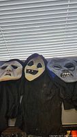 Three Halloween latex masks with black fabric hoods displayed horizontally on surface under window blinds. Each mask depicts a different ghastly face.
