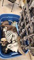 View of blue storage bin with Halloween skeleton parts including skull and torso, decorative fence panels leaning upright, and a boxed Halloween item inside the bin.