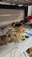 Photo of a blue velvet and gold crown with multicolored jewels, blonde wig, white gloves, and other costume accessories.