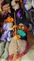 Close-up showing a witch doll holding a broomstick with a burlap body and black hat, orange hair and detailed hands.