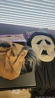 Two Halloween masks shown side by side on a surface near a window, one tan with pointed ears and gray hair, the other white screaming face with black fabric.