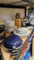 Multiple stacks of white ceramic plates, wooden block with knives and utensils, blue frying pan with detachable handle visible