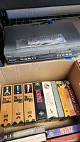 Photo showing Panasonic VHS/DVD player on top of a box with a remote control in plastic bag and VHS movies in box beneath, including The Godfather series, Scarface, Silence of the Lambs, Witness, The Untouchables, and The Joy Luck Club.