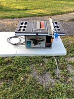 Front side view of a Shop Craft table saw with cord coiled next to it on a white folding table outdoors