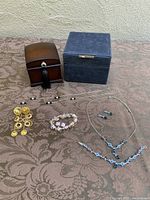 Photo showing two jewelry cases, one wood Bombay style box with tassel and one blue vegan leather square case, alongside assorted costume jewelry including blue crystal drop necklace, earrings, bracelet set, yellow gold-tone dangle earring, pink floral bracelet, and blue stud earrings.