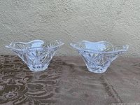 Two clear crystal bowls with scalloped edges set on a textured brown surface, showing the profile and intricate cuts in the glass.