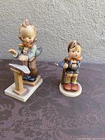 Both Band Leader and Little Hiker Hummel figurines together on a patterned cloth surface with a textured wall background.