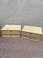 Two wooden storage boxes side by side on a fabric surface in front of a gray wall. Each box has three drawers with circular finger holes for opening.