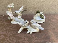 Three porcelain Otagiri figurines displayed on brown cloth: hummingbird on flowering branch, two birds on a branch, and frog music box