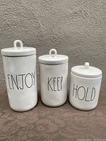 Three white ceramic Rae Dunn canisters of varying sizes labeled ENJOY, KEEP, and HOLD with lids and loop handles.