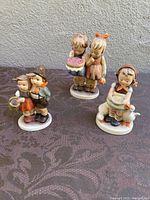 Photo of all three vintage Hummel porcelain figurines together on a dark patterned cloth background.