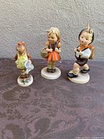 Front view of three Hummel figurines on table: Little Gardener, School Girl, School Boy.