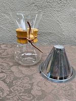 Full view of glass coffee carafe with wooden collar and leather tie alongside stainless steel conical coffee filter, set on textured fabric surface.