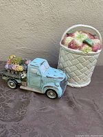 Co-located photo showing the flower basket cookie jar and flower shop truck figurine together on a dark patterned surface.