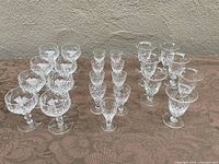 Set of 18 crystal glasses arranged on a patterned cloth, clear with diamond and cross-cut designs, includes multiple sizes and types.