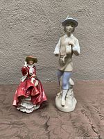 Two standing porcelain figurines together, Royal Doulton on left in red dress, Miguel Requena on right in light clothing with basket