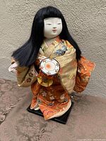 Front view of a 13-inch Japanese doll dressed in a patterned orange and gold kimono holding a small drum fixed to a handle.