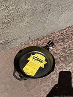 Photo showing the Lodge cast iron frying pan on a textured surface with original yellow label inside the pan.
