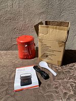 Photo of the red rice cooker with its original cardboard box, power cord, instruction manual and rice paddle shown on a table outdoors.
