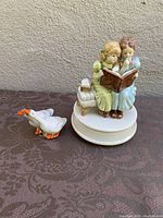 Wide shot showing the porcelain music box of two girls reading a book and a small goose figurine on a patterned cloth background.