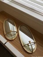 Two oval mirrored wall mount candle holders on wooden surface showing gold-tone metal frames and candle cups