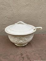 White ceramic soup tureen with lid and ladle, embossed olive branch design, on patterned surface against beige wall, front angle