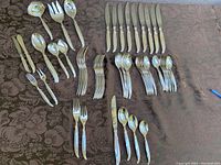 Overview of all flatware pieces including knives, forks, spoons, serving utensils laid out on dark brown patterned cloth.