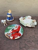 Full view of all items on display including the angel music box, lamb figurine, Coca-Cola Santa plate, and Fitz & Floyd Santa plate on a patterned cloth.