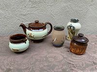 The full lot of five pottery items arranged outdoors on a brown patterned cloth showing teapot, creamer, two vases, and small lidded pot