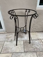 Wrought iron pub table base standing on tiled floor against wall, showing side view with four legs and scroll decoration.