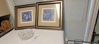 Two taupe wood framed artworks featuring abstract compositions of pears and grapes displayed on a white cloth surface under window blinds.