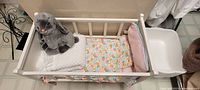 White wooden baby doll cradle holding blankets and grey stuffed animal, seen from above.