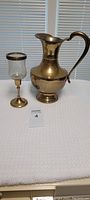 Photo showing large brass jug and one brass and glass candle holder on a white surface in front of window blinds.
