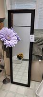 Full view of tall rectangular mirror with dark brown frame next to the lilac flower in standing holder, showing size and condition in living room setting.