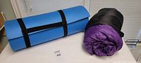 Blue foam sleeping mat rolled and strapped beside a rolled purple sleeping bag in a black sack on a white surface