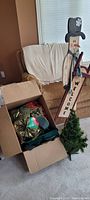 Photo showing assorted large red and green Christmas bows in box along with the small Christmas tree and wooden snowman welcome sign leaning against chair.