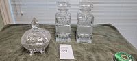 Photo showing two clear crystal decanters with geometric cut designs and rectangular stoppers, alongside a footed round clear crystal candy dish with lid and ornate pattern.