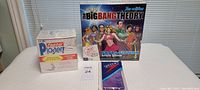 Three items on a white padded surface: The Big Bang Theory trivia game box, plaster kit box labeled 'Elite Faster Plaster!', and a purple Speedo bathing cap in a plastic package.