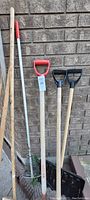 Garden tools including 2 red-handled shovels, 2 black plastic snow shovels, rakes with metal heads, and wooden-handled brooms stacked against a brick wall.