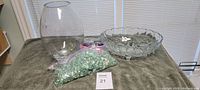 Overview of the lot showing the glass vase, a glass bowl, and packs of glass beads on a towel with window blinds background.