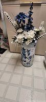 Photo showing the full vase standing on floor, displaying tall blue and white silk flowers arranged inside a blue and white ceramic vase with floral details.