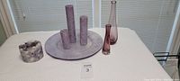 Wide view of lot on white textured surface with items arranged: three candles on large lilac plate, two vases, lilac and white speckled candle holder.