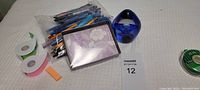 Photo showing a bag of assorted pens, pencils, crayons, a box of tissues or wipes, rolls of colored sticky tape, and a blue glass paperweight on a light surface.