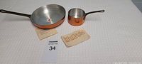 Two mini copper pans set laid on a white textured surface with tags visible. One is a 6-inch fry pan and the other is a 3-inch small pot. Both have copper exteriors and stainless steel interiors with black handles.