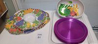 Two large fruit print platters stacked with a plastic segmented serving bowl with fruit and vegetable print on top, all shown on a clear plastic tray.