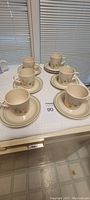Six coffee/tea cups placed on matching saucers, all arranged on a surface against a window blind background, showing overall set condition and design.