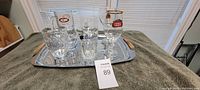 Silver metal tray on table with five glass beer steins and mugs with brand logos, close to window with blinds.