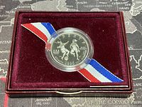 Silver-colored commemorative half dollar coin in clear capsule, situated in burgundy velvet interior with red, white, and blue ribbon attached.