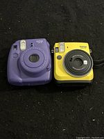 Purple Instax Mini 8 and yellow Instax Mini 70 instant cameras side by side on black surface.