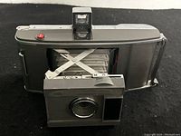 Front view of vintage Polaroid J66 Land Camera with extended bellows and lens panel open, showing the red shutter button and pop-up viewfinder.